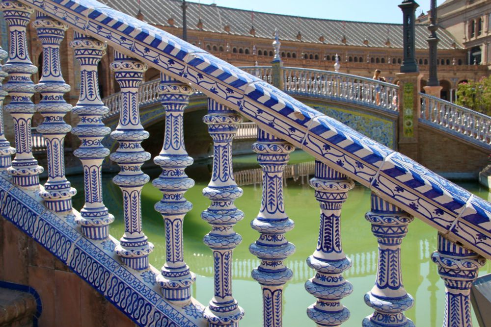 part of a white and blue ceiling covered with symbols and patterns (eyes, stripes, floral lines), green water ist visible through the gaps between the single parts, and a rounded bridge with a different ceiling and big mosaiques in yellow, white and blue on parts of it, in the background parts of a big palace in brown with little arched windows 