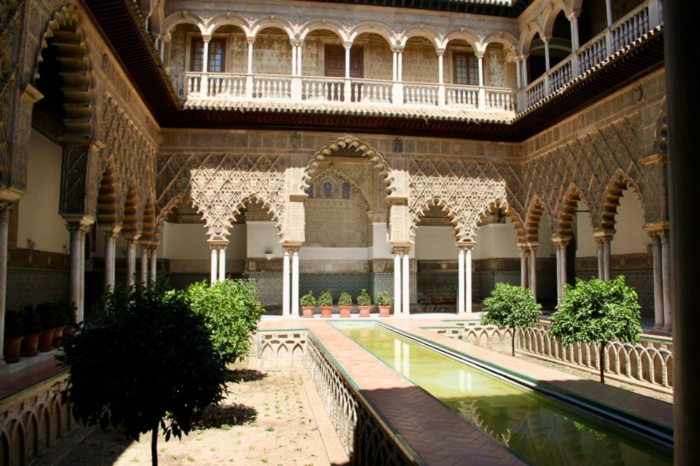 The patio of the Alcázar, three parts of the building are visible, a lot of arches covered by ornaments, small columns, bigger arches, a second floor with more arches and some doors, small citrus trees in two parallel rows, in the same parallel row a rectangular pool with gree water, ceilings framing the whole garden, on some of them plants in flowerpots.
