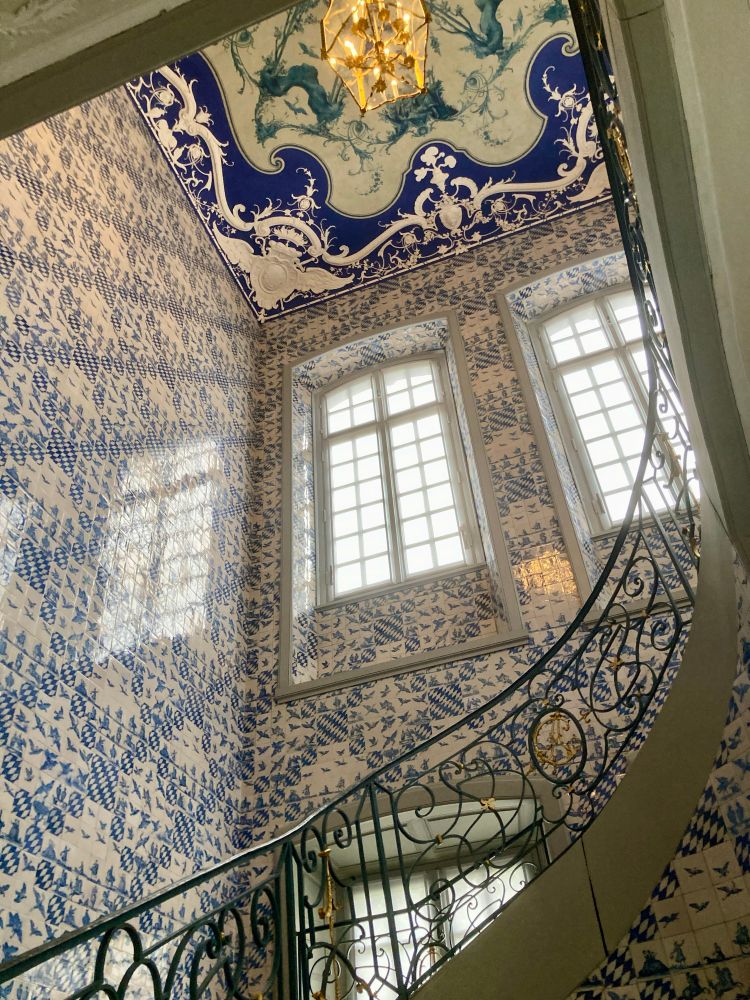 Staircase of a palace, the walls are decorated all over with blue and white mosaics made of a shining ceramic stone. Part of a black curved  wrought iron railing with ornaments. The staircase is illuminated by the sunlight shining through big white framed glass windows, one is reflected on the wall. The ceiling is also decorated with black and white and golden patterns like a lace tablecloth and a golden chandelier ion the center.