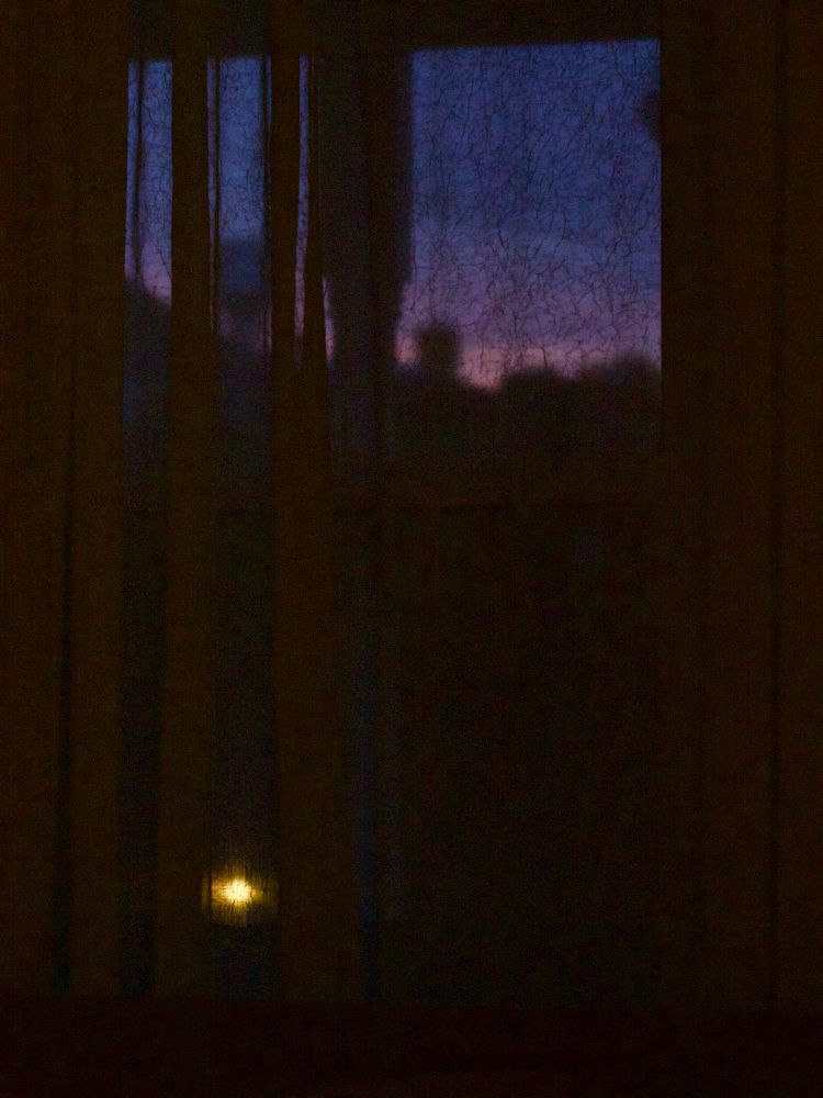 A darkening, deep blue sky is seen over rooftops through a gauzy curtain. A yellow light in the lower left corner floats in darkness