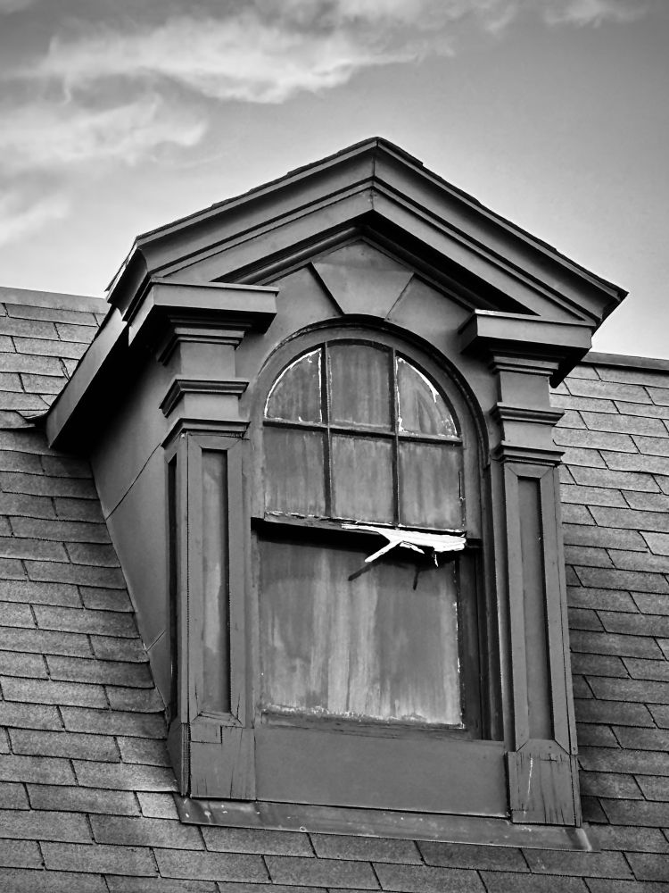 Black and white photo of a dormer showing weathering such as cracked and split wood, and evidence of fading black paint on the inside of the window panes.