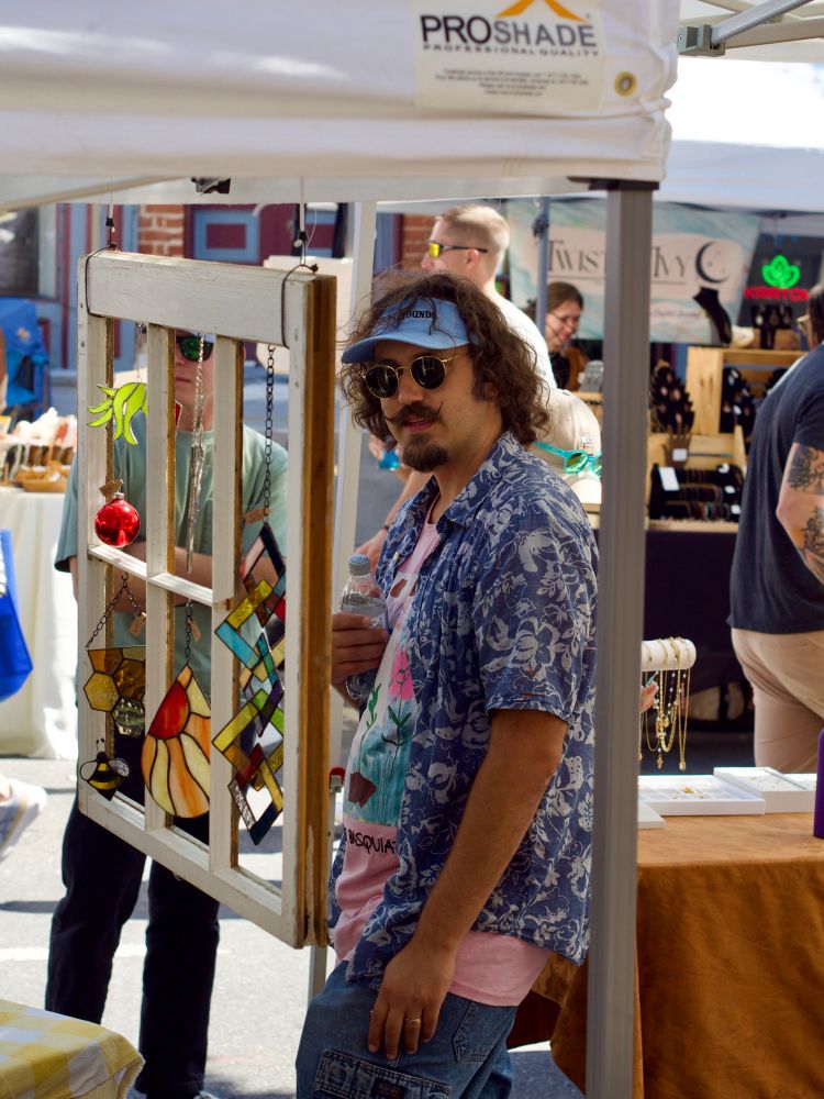 A man wearing a colorful Hawaiian shirt and a sunshade sports cool sunglasses while he looks at glass art at a street festival