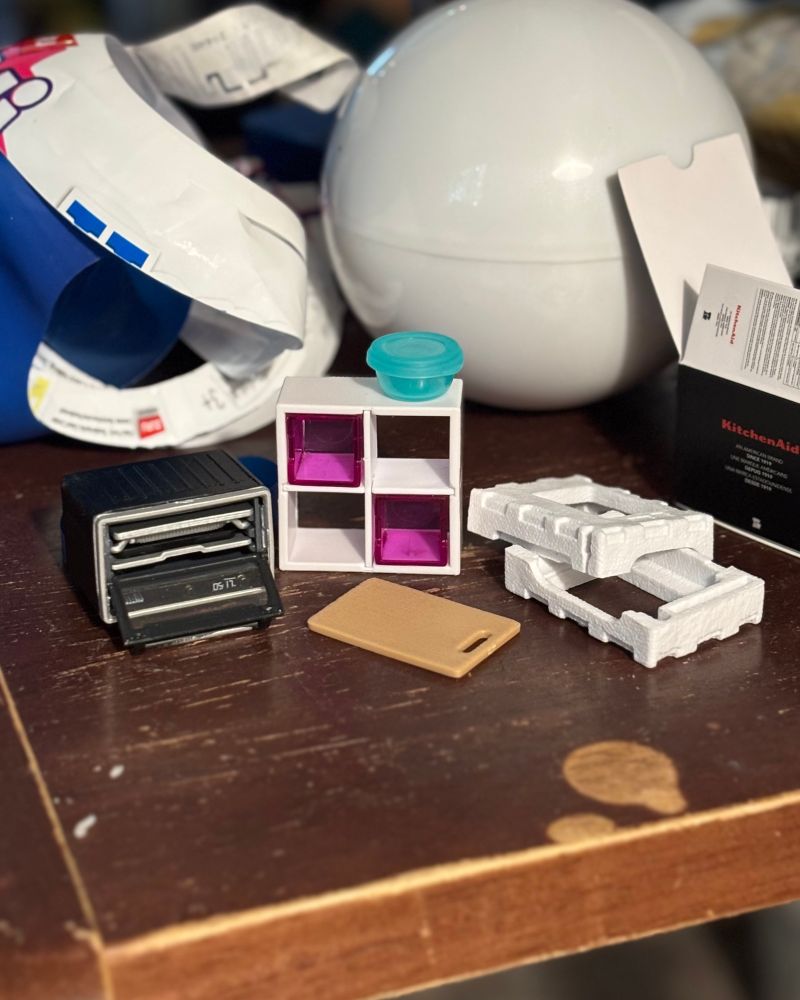 a tiny black toaster oven that is less than an inch tall, a tiny white shelf with tiny pinky-purple cubes in two of the slots. a tiny teal food container that is about the size of a fingernail rests on top of the tiny cube shelf. in front of the shelf is a tiny light brown cutting board, about an inch wide. On the right is tiny packing foam things for the tiny toaster oven, and a tiny KitchenAid-branded box for the tiny toaster oven. behind it is the good-awful amount of plastic i had to unwrap to get to the tiny kitchen items. i used the word “tiny” in this description twelve times. 