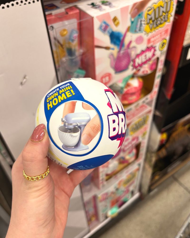 a hand holding a Mini Brand ball, with a tiny KitchenAid mixer pictured on it, with the words “CREATE YOUR OWN MINI HOME!”. behind it is a stand of colorful boxes that have other miniverse bullshit in them. 