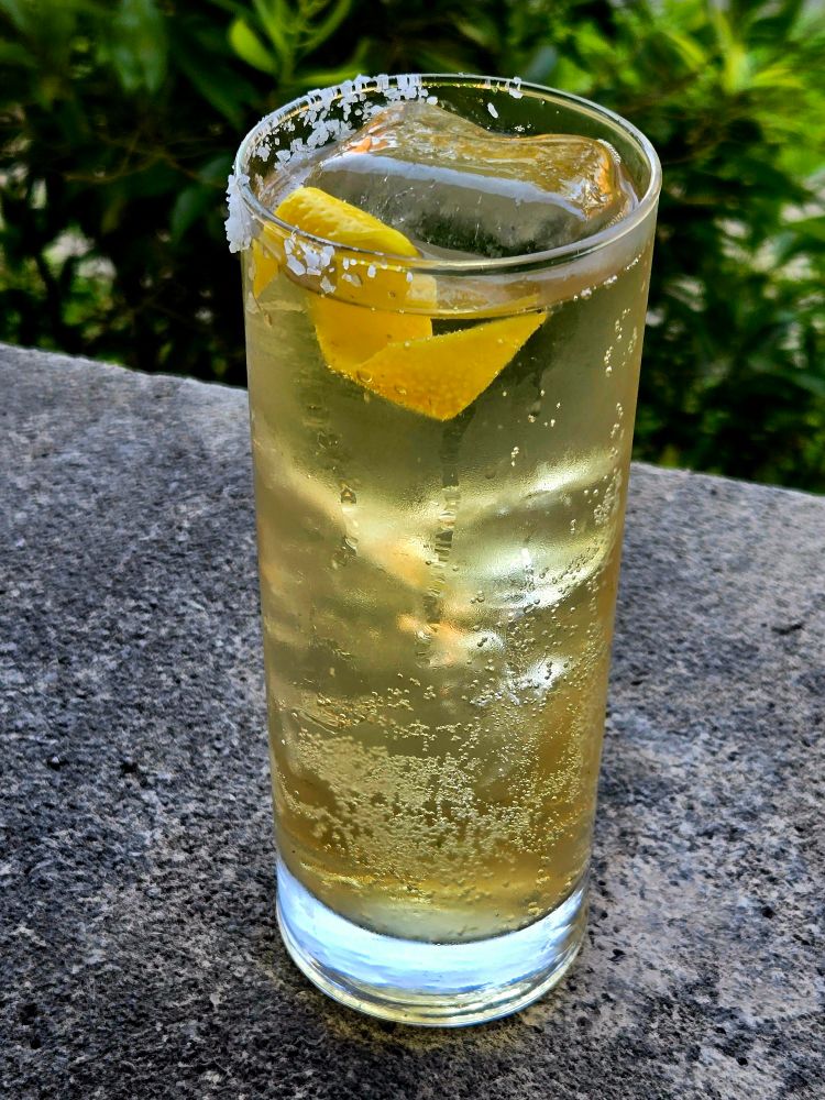 A Scotch highball with lime cordial and lemon garnish in a highball sitting on stone. 