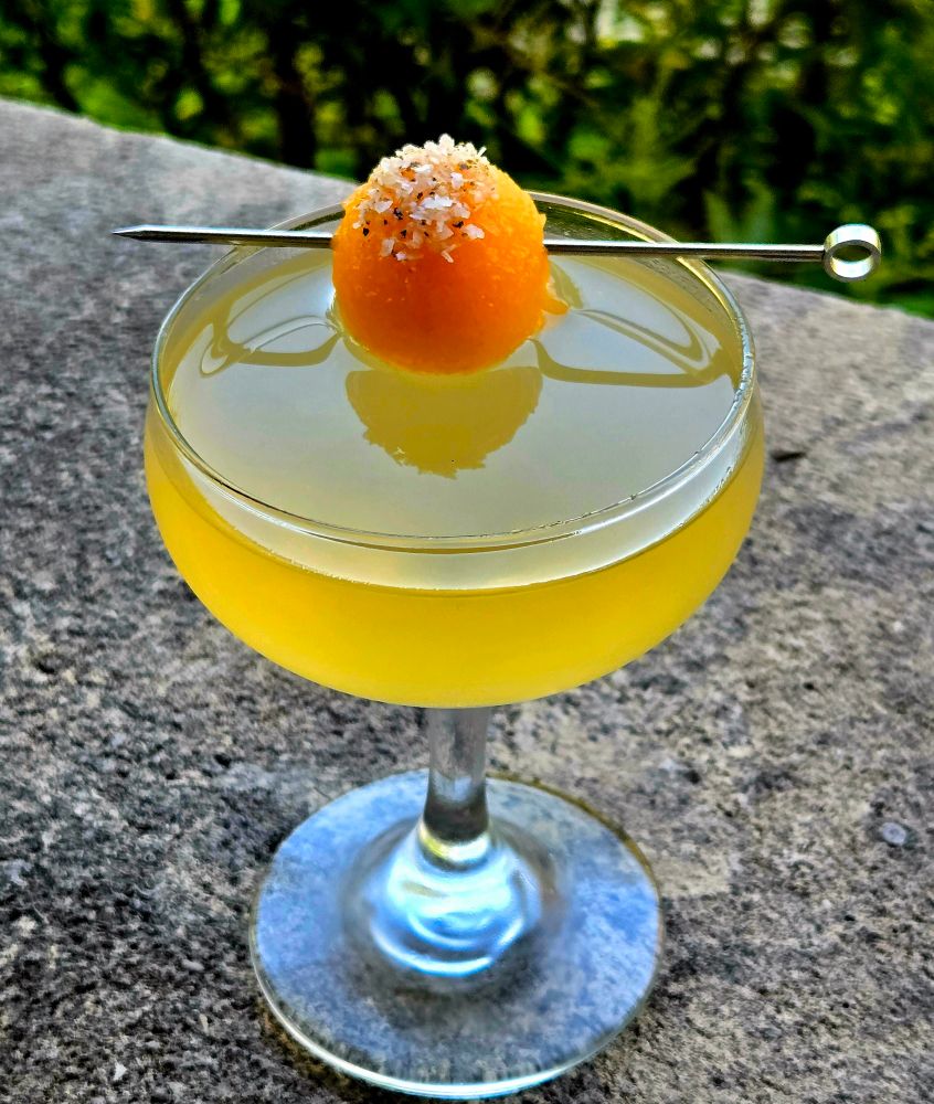A cocktail of tequila, Cantaloupe, pink peepercorn, and lime in a coupe glass atop stone. 