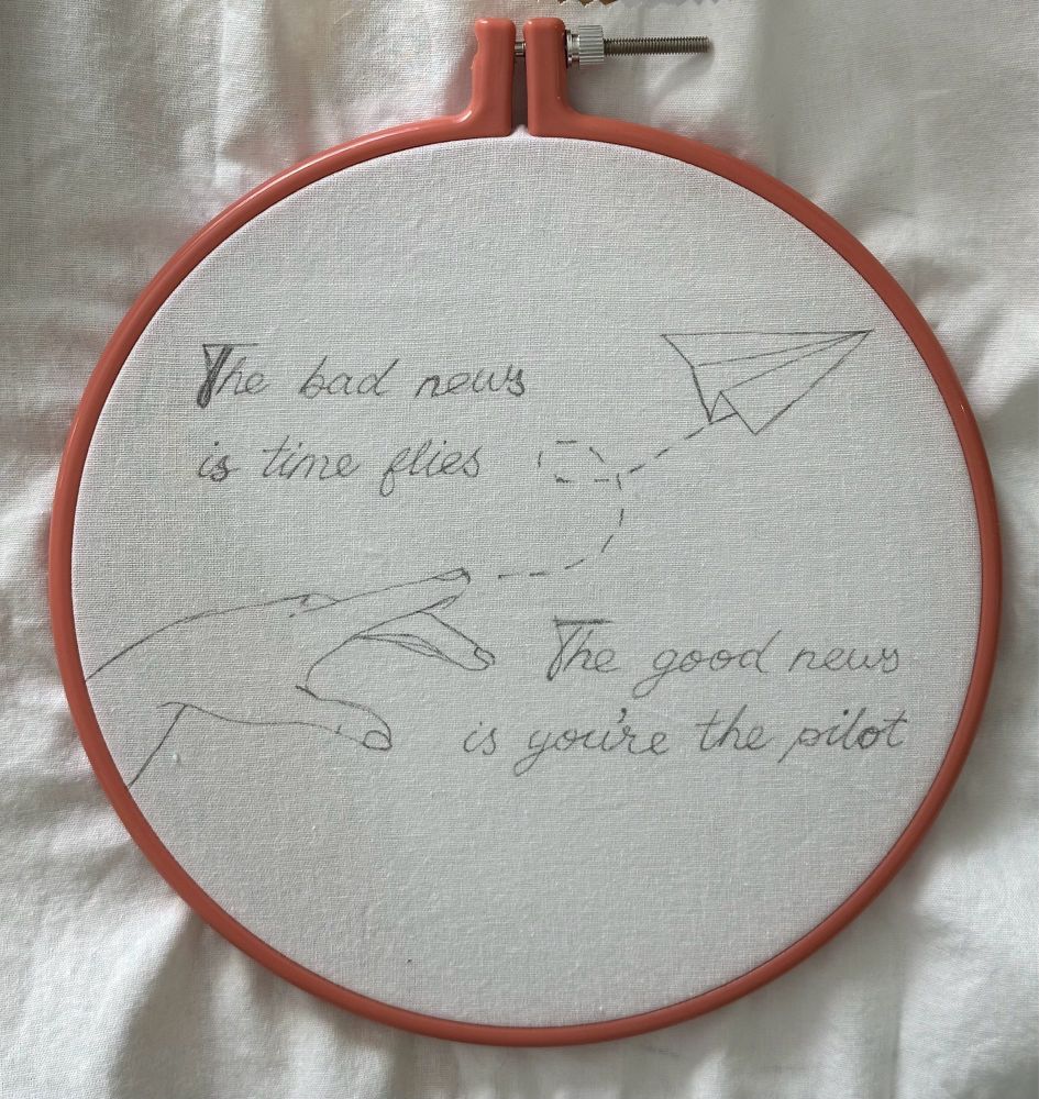 White fabric in a dusky pink embroidery hoop. Traced onto the fabric in black pen is a line drawing of a paper airplane (top right) thrown by a hand (lower left) and the looping path the airplane took. Above the hand are the words “The bad news is time flies” and to the right of the hand are the words “The good news is you’re the pilot”.