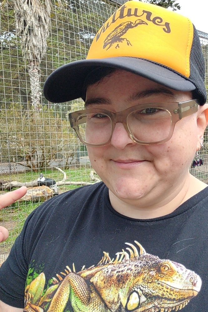 a selfie of me standing in front of the condor exhibit pointing to the seated condor in the background. i am wearing a bright yellow hat with a vulture and the word "vultures" on it. i'm wearing a black shirt with a realistic green iguana on it.