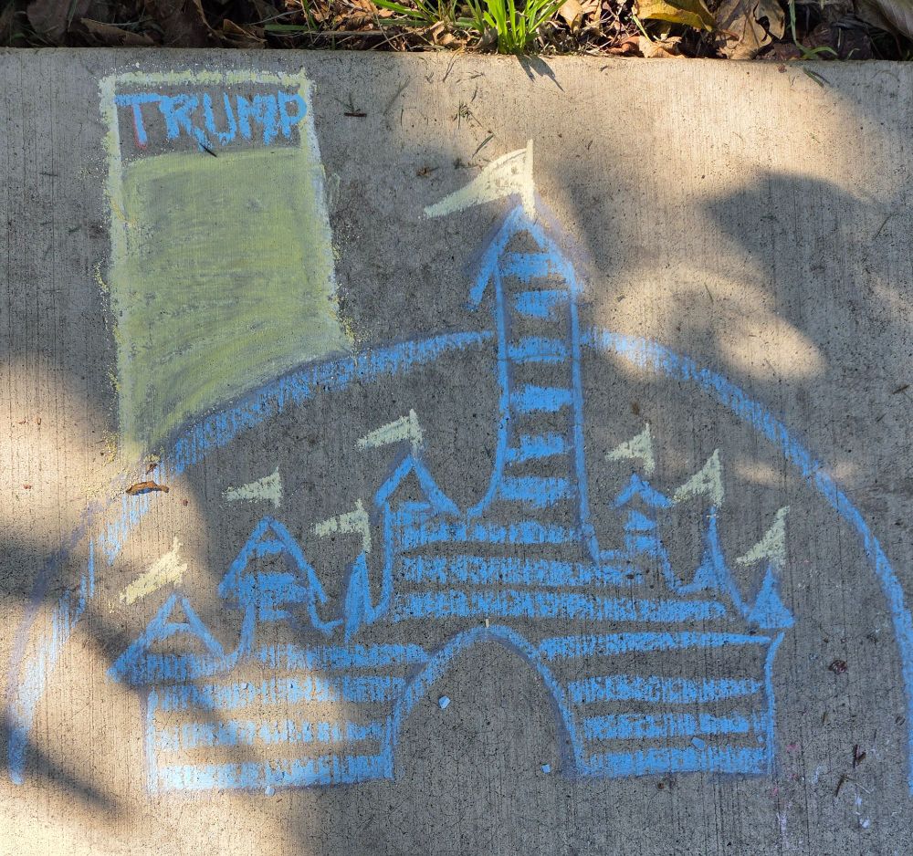  Chalk drawing on sidewalk in a park. Disney films logo of a blue castle with flag-topped turrets and a half circle arching over it.  Looming above the logo is a golden skyscraper with "TRUMP" at the top of it. 
