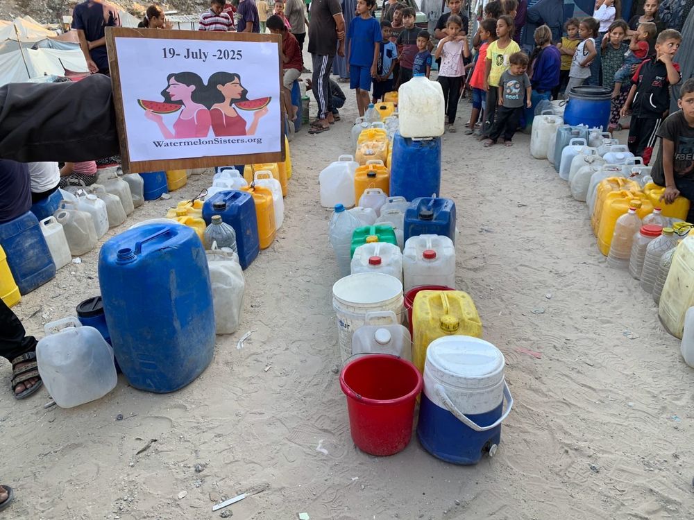 A photo of a sign saying 19 July 2025 with multiple carriers of water surrounded by people