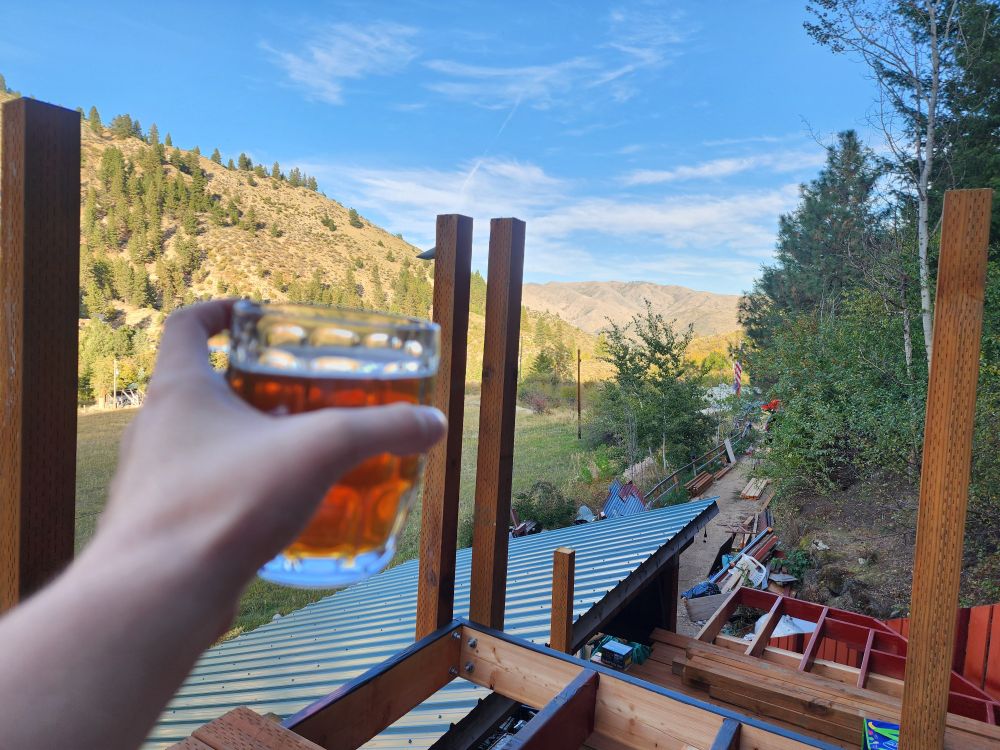 Someone extends their hand holding a beer mug looking out over an incomplete deck and a canyon at late day. 