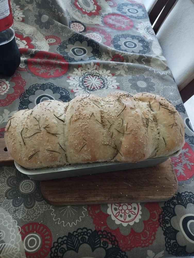 Pão assado dentro de uma forma de metal. O pão possui sementes de gergelim e folhas de alecrim em cima.