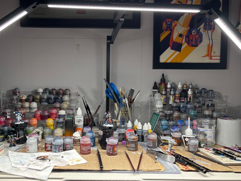 Messy desk covered with brushes, various paint pots, cardboard and Warhammer minis in various stages of being painted. 