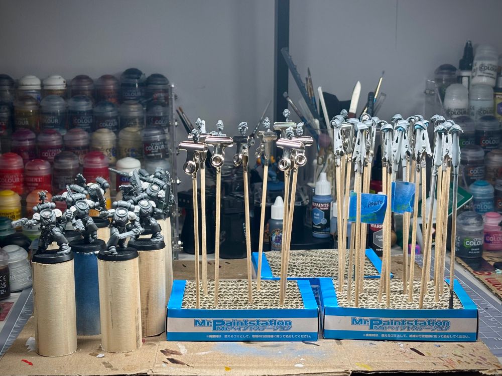 Desk with paint pots in the background. In the foreground are minis in a broken down state of subassembly ready to be primed. 