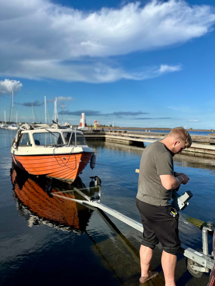 A man stands barefoot in shallow water next to a boat trailer, adjusting equipment as a small orange and white motorboat floats behind him. The scene takes place at a marina on a clear, sunny day, with docks, sailboats, and a lighthouse visible in the background under a bright blue sky with scattered clouds.