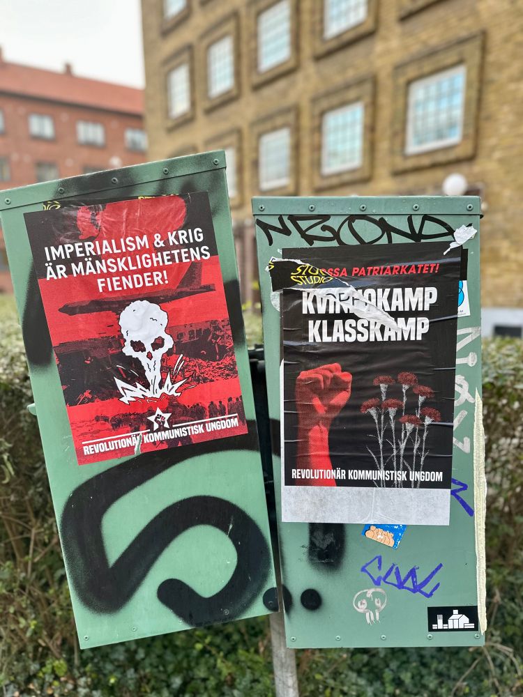 A photo shows two posters attached to a green utility box outdoors, with a brick building and hedge in the background. The left poster is red and black, featuring a skull and explosions, with the Swedish text: “IMPERIALISM & KRIG ÄR MÄNSKLIGHETENS FIENDER! REVOLUTIONÄR KOMMUNISTISK UNGDOM” (“Imperialism & war are humanity’s enemies! Revolutionary Communist Youth”). The right poster is black and red, showing a raised red fist and red carnations, with the Swedish text: “KVINNOKAMP KLASSKAMP REVOLUTIONÄR KOMMUNISTISK UNGDOM” (“Women’s struggle is class struggle. Revolutionary Communist Youth”). Both posters promote a left-wing political message.