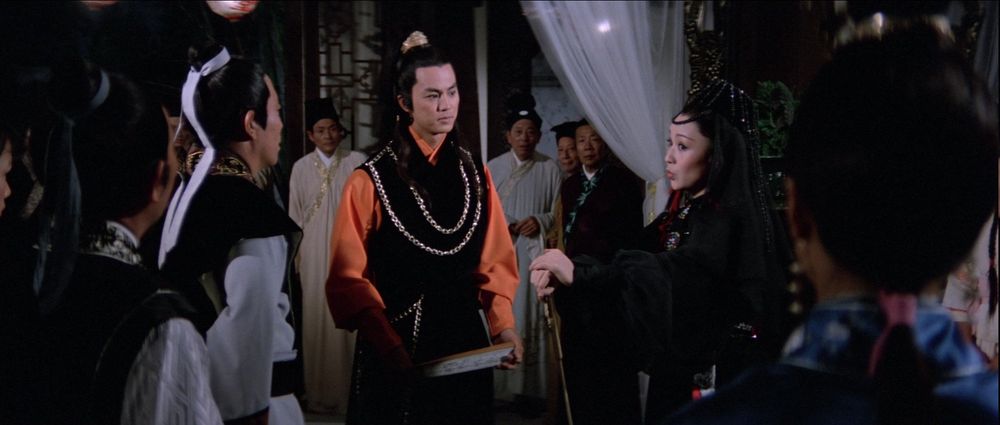Ti Lung in a striking orange and black costume, talking with Li Ching in an exotic costume, in an ornate gambling den.