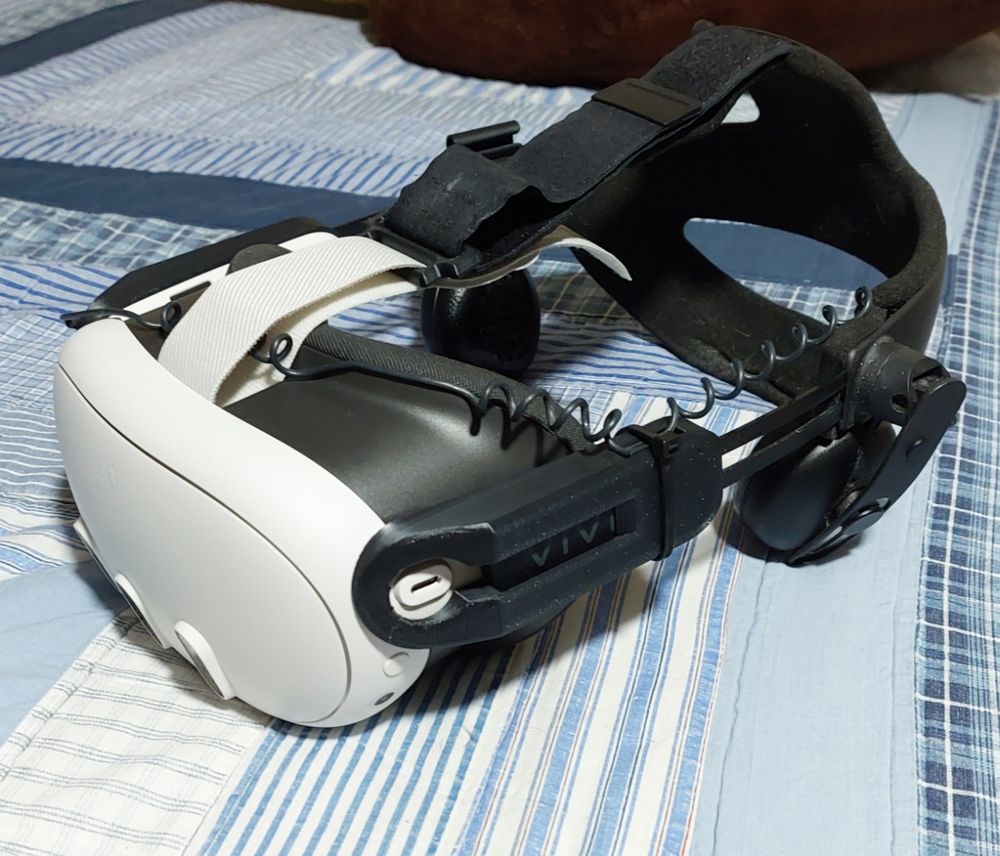 A Quest 3 modded with a HTC Vive Deluxe head strap. The head strap is attached to the headset via 3D printed attachments on the sides and on the loop of the strap.