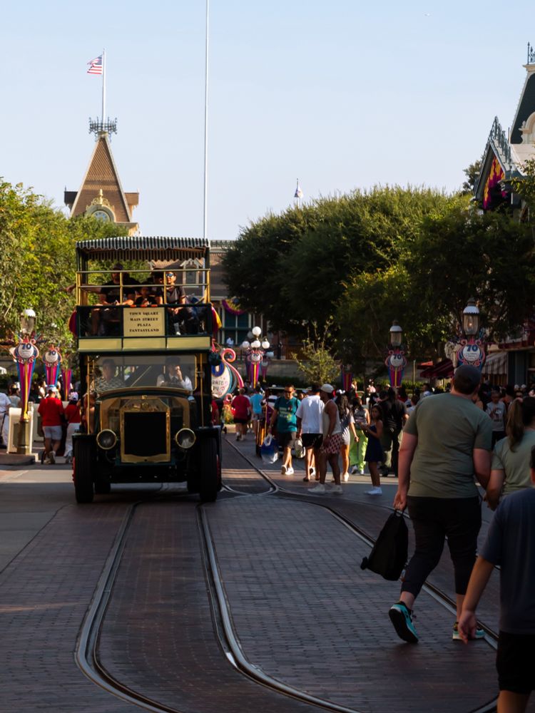 The Omnibus, partially obscured in shadow, drives up Main Street USA with the railroad station in the background as people clear out of the way on the street.