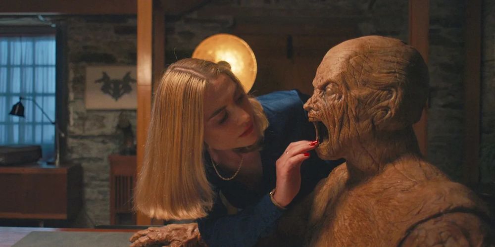 a still from the movie "oddity" (2024) featuring a blonde woman with a long bob peering into the mouth of a horrifying wooden mannequin with its faces fixed in a scream. her fingertips brush its mouth, her nails painted blood red. 