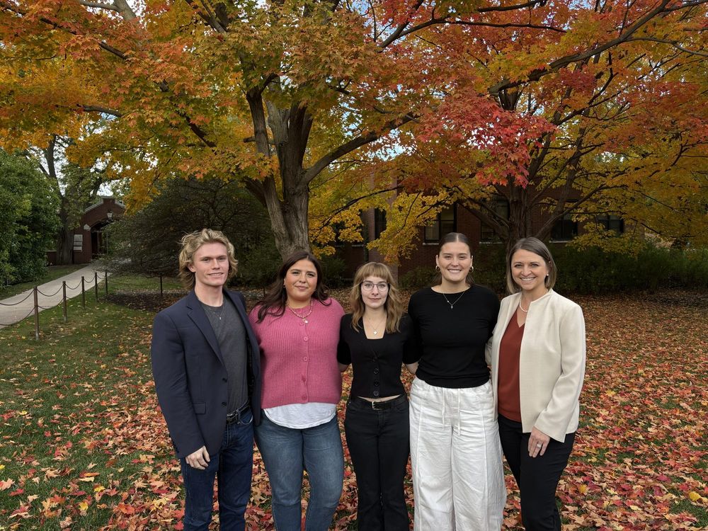 5 adults standing together and smiling in front of a tree with colorful leaves in hues of yellow and red.