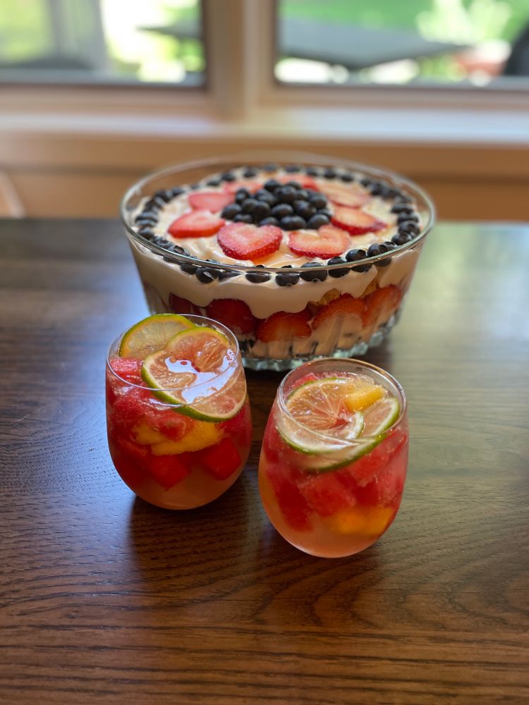 Two glasses of summer white sangria with fresh cold watermelon cubes, fresh pineapple pieces, thinly-sliced fresh lime over ice in front of a trifle dessert bowl with fresh strawberries, blueberries, layers of angel food cake, and white chocolate pudding mixed with whipped topping.