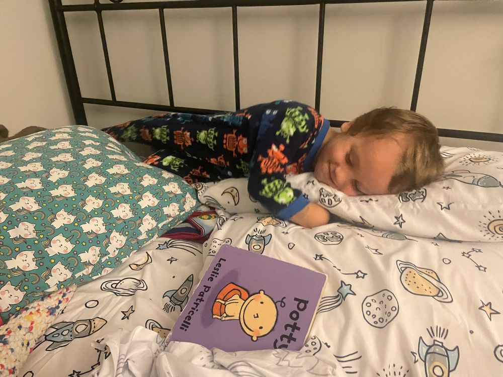 A preschooler in robot jammies on rocket sheets with a Capricorn pillow. He’s smiling and looks very sweet