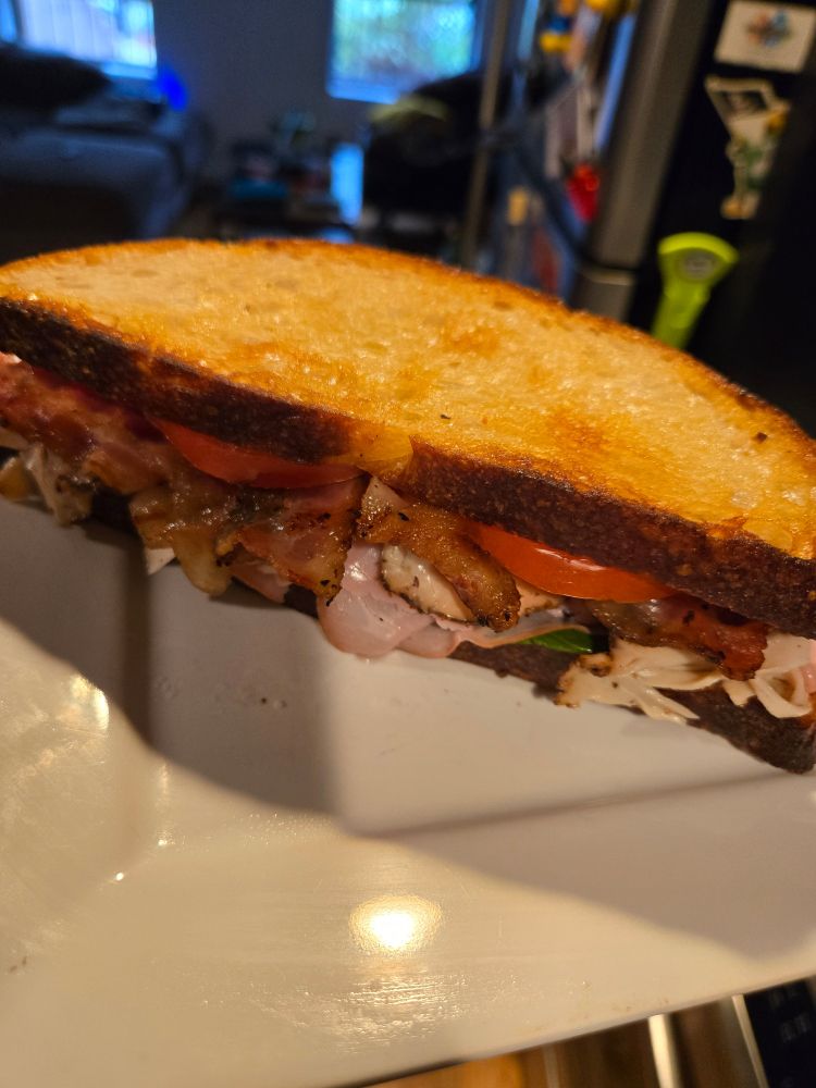 A white plate holding a sandwich of toasted sourdough bread, tomato, basil, pepper bacon, turkey pastrami, honey ham, a light spread of Dijon on one side and Wegmans triple cream brie on the other