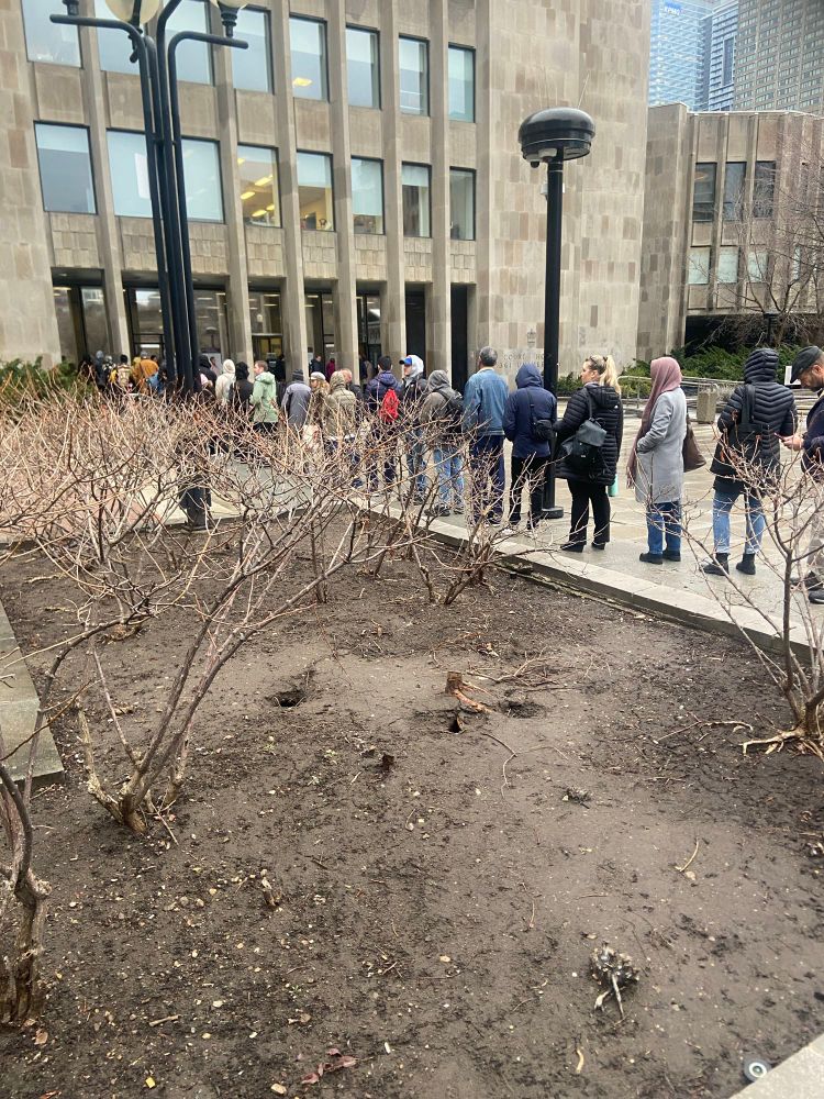 A long line of damp people standing in a grey morning outside a grey building. 