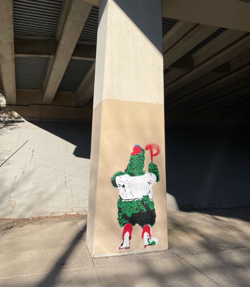 Banksy style graffiti of the Phillie Phanatic spray painting the Phillies logo on a highway support column 