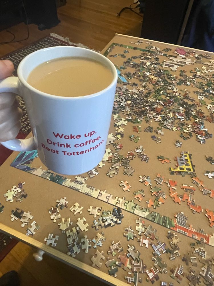 A white mug of tea that has “Wake up. Drink coffee. Beat Tottenham.” written in red on it held in front of a just-started jigsaw puzzle. 