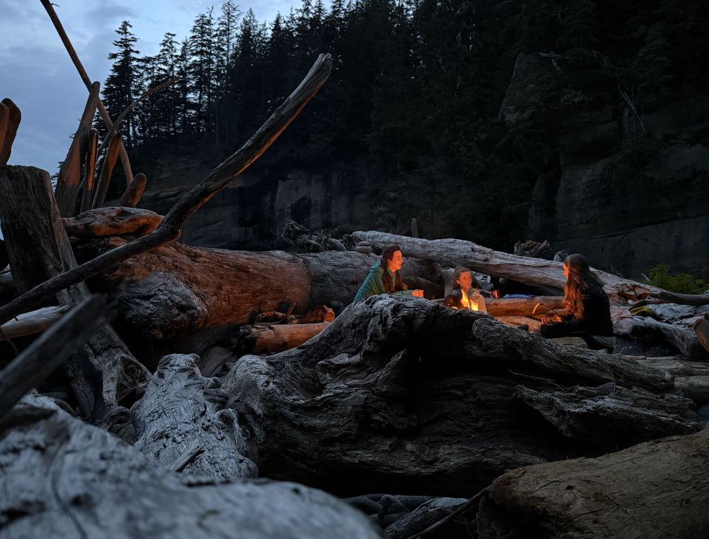 Photo of three women sitting around a fire. The picture is framed and the fire is mostly hidden by large pieces of drift wood. 