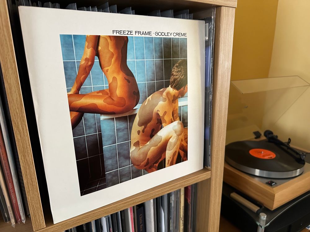Album cover for Godley & Creme’s  Freeze Frame.  Cover depicts a man and woman in silhouette in a blue tiled bathroom.  Their skin has a vitiligo like camouflage effect all over.