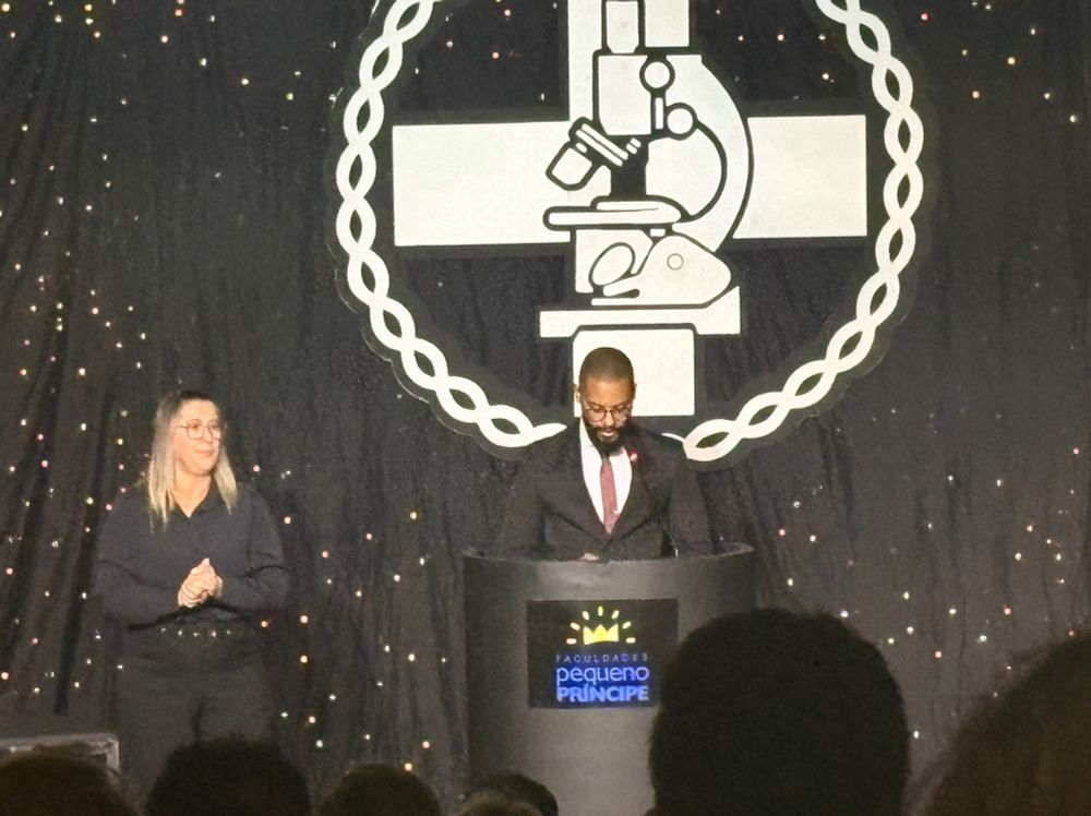 Foto do palco, com fundo preto e símbolo de biomedicina em branco. Diante do símbolo, mestre de cerimônia diante do púlpito e intérprete de libras a sua esquerda.