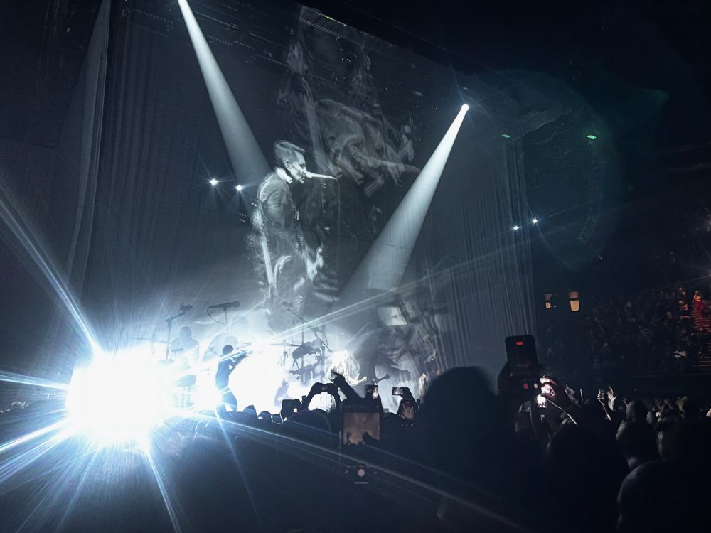 A band playing with bright white lighting silhouetting them, live images projected on layers of scrim. 