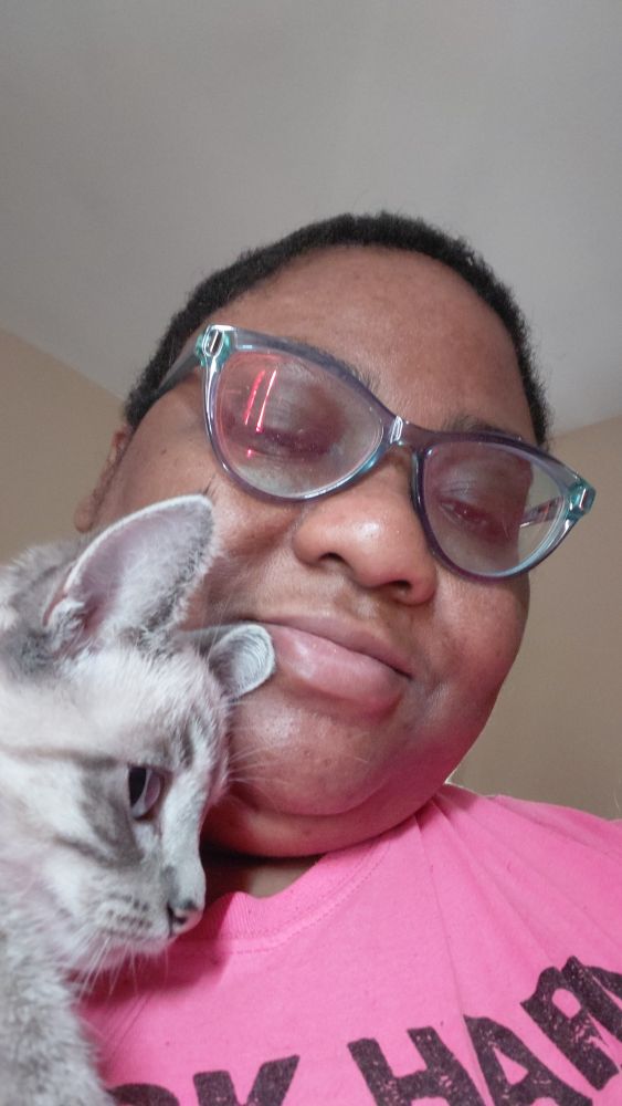 Darunia, a Black non-binary person with green glasses and a pink shirt, is holding a color point cat named Crystal. They are smiling at the camera, while Crystal is, as always, judging her owner.