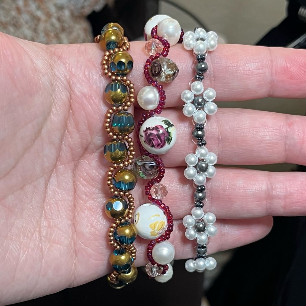 Three handmade bracelets; the first is made of teal and bronze faceted beads with small bronze seed beads winding back and forth around them. The second is another mix of large ceramic beads with dark pink floral and faceted glass beads, with dark red/purple seed beads winding between them. The third is a daisy chain style with round white pearls in the shape of flower petals and dark grey pearls in the center. 