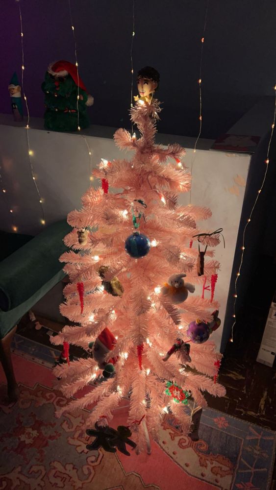 A small pink faux christmas tree decorated with lights and ornaments and topped with a Kurtis Conner Youtooz figure