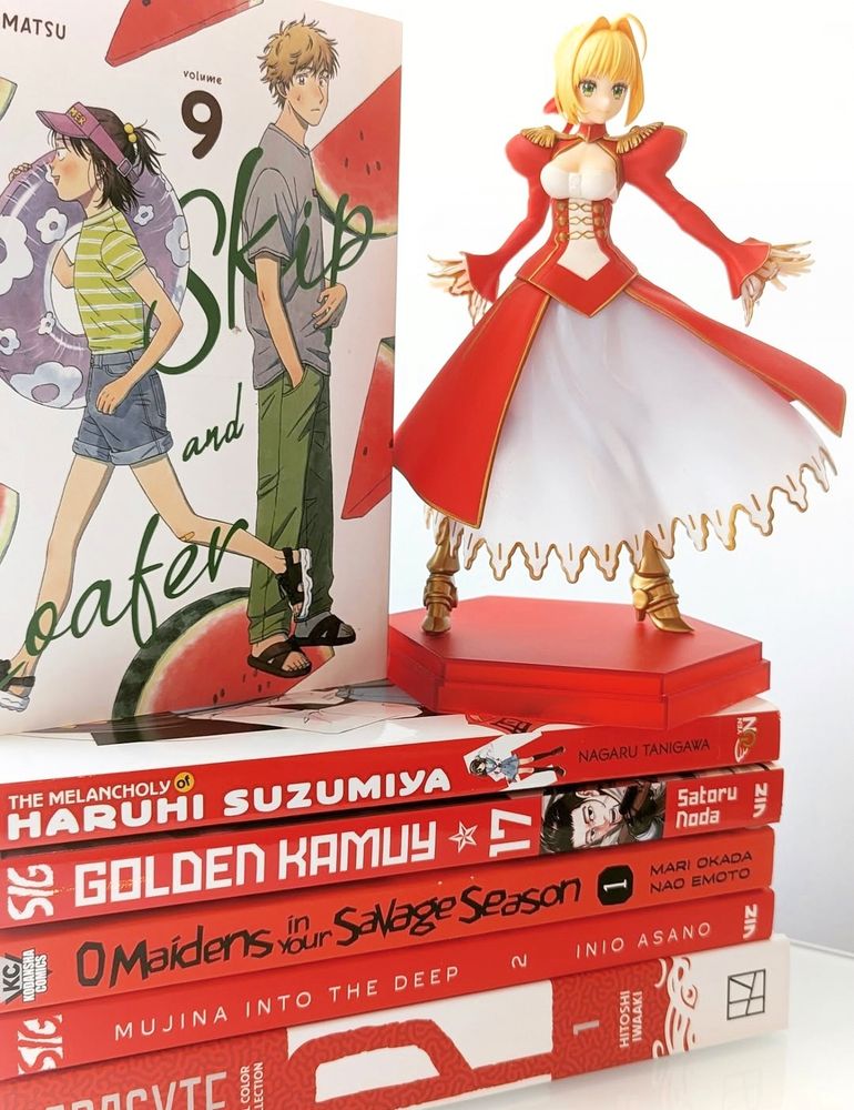 A stack of bright red manga, with the cover of Skil and Loafer volume 9 atop them and paired with a pop up parade figure of Nero Claudius.