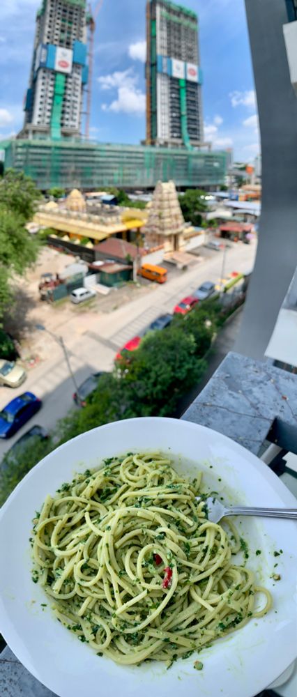A bowl of spaghetti with pesto sits on a balcony railing. In the background, on the street, is a Hindu temple, with two under-construction high-rises behind it, backdropped by a bright sky with puffy clouds.