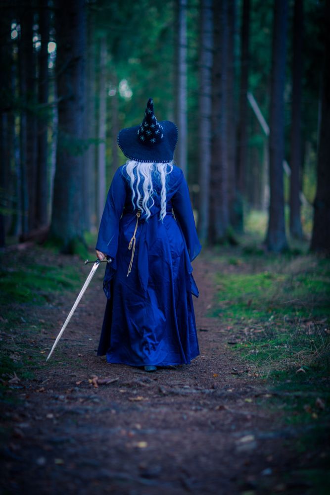 Eine Person in einem langen, dunkelblauen Hexenkleid und spitzem Hut, mit langem weißem Haar, steht von hinten auf einem Waldweg und hält ein Schwert in der rechten Hand. Der Weg führt tief in einen dichten Nadelwald.