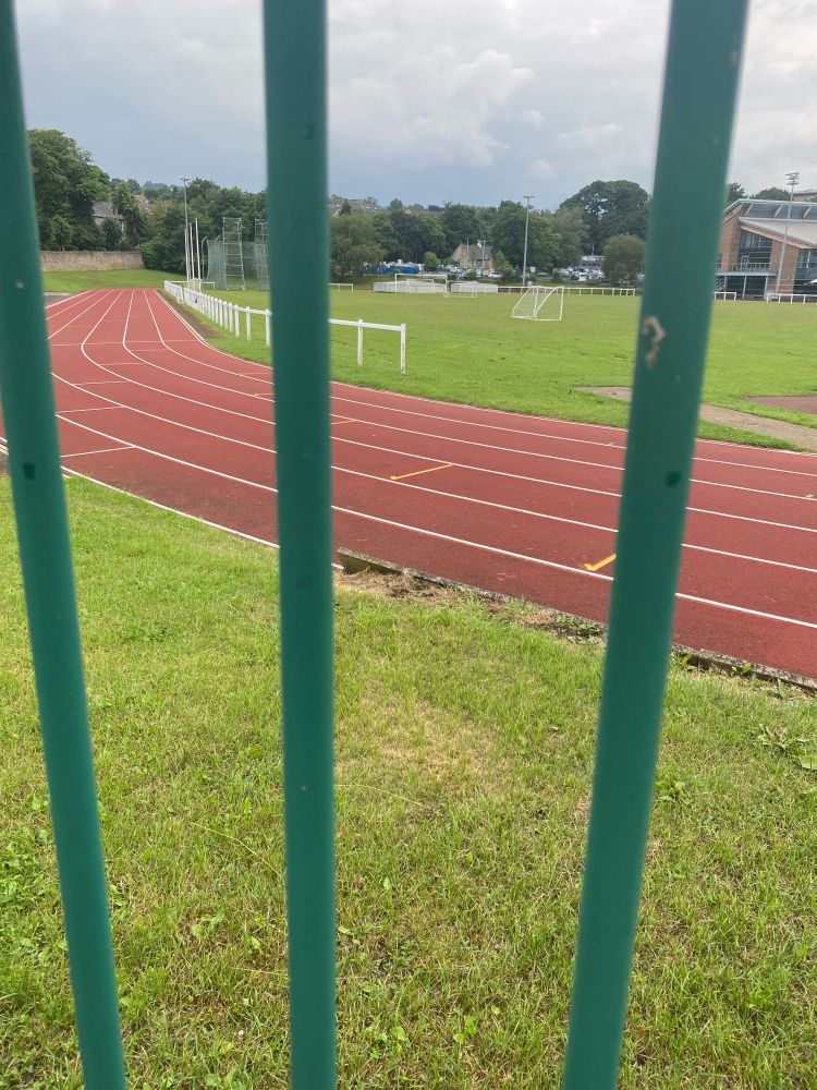 A view through three horizontal metal bars. Beyond them, the bend of a running track and a patch of grass which looks like one end of a football pitch. Also some sort of pavilion building. 