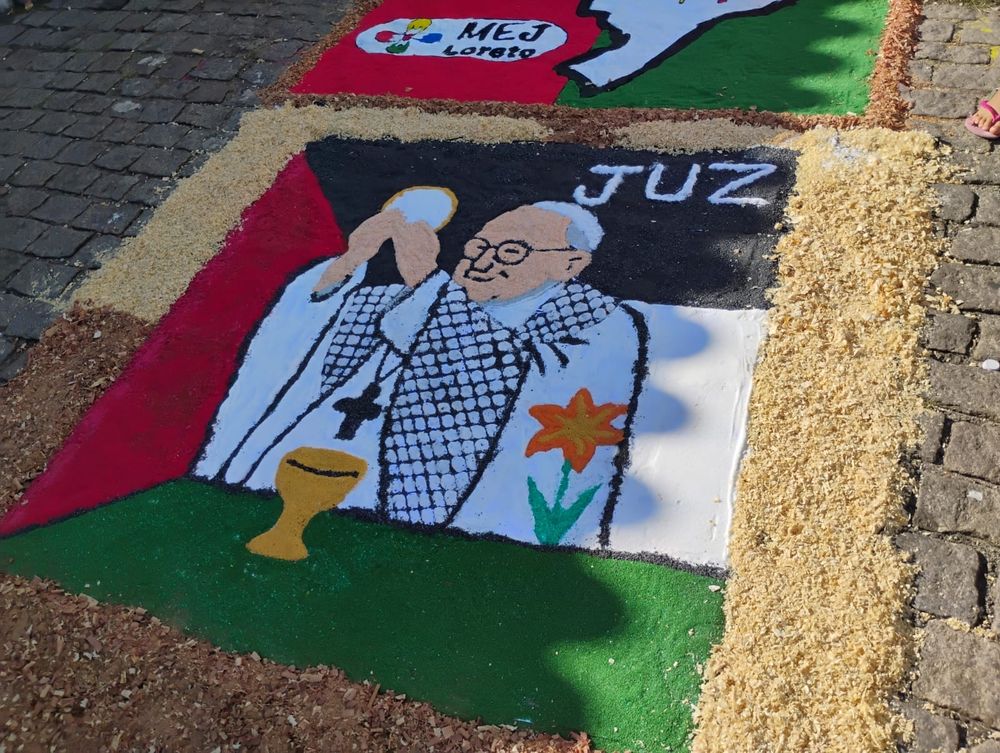 Tapete de sal de Corpus Christi num chão de paralelepípedos. O tapete tem a imagem do Papa Francisco no momento da consagração (do rito católico) com a hóstia nas mãos. Na sua frente podemos ver o cálice e do lado esquerdo do papa, um lírio do campo, representando santo Antonio Maria Zaccaria. O papa usa vestes brancas e um keffiyeh sobre os ombros. No canto superior direito da imagem tem-se a grafia "JUZ" - representando o nome do grupo, e o fundo da imagem é a bandeira da Palestina. 