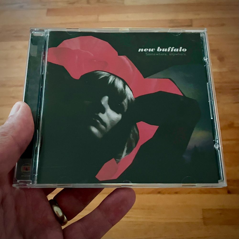 A hand holding a CD case over a wood floor. The cover art is a black & white, shoulders-up shot of a woman in stark shadow, holding her hands behind her head. She is framed by a red cut-out shape, possibly a flower, and a greenish, shadowy background.