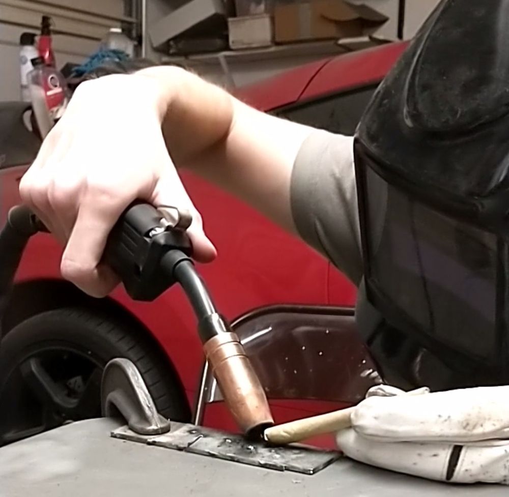 me with a welding mask on holding a MIG welding torch against a sheet of metal with a joint inbetween