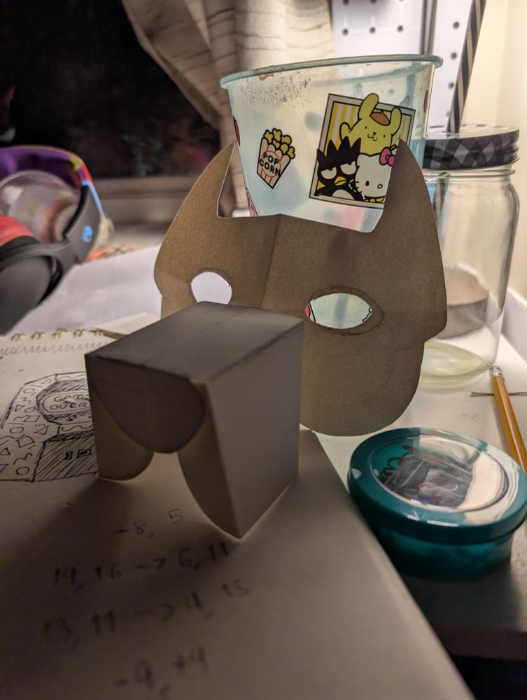 A paper template for a dog mask, based off a cane corso. It's a simplistic template. Sitting on a sketchbook, leaned against a hello kitty cup.