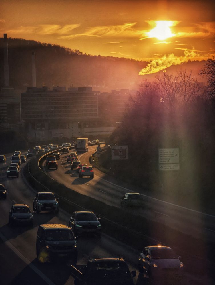 Das Bild zeigt eine Schnellstraße bei Sonnenuntergang. Mehrere Spuren der Straße sind mit dichtem Verkehr gefüllt. Zahlreiche Autos sind zu sehen, viele von ihnen in einem leichten Stau oder in langsamer Bewegung. 
Die tief stehende Sonne wirft lange Schatten und erzeugt starke Lichtreflexe (Lens Flares) auf der Fahrbahn und durch das Bild. Über der Straße erstreckt sich eine bewaldete Anhöhe. Am Fuße des Hügels, hinter der Schnellstraße ist eine größere Gebäudestruktur zu erkennen. Die Beleuchtung ist warm und dramatisch. Der Himmel glüht in Orange- und Gelbtönen,