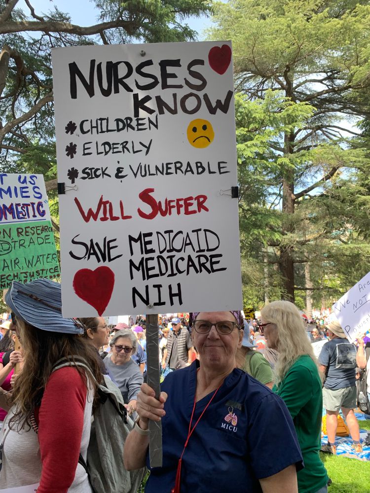 A nurse in scrubs with a sign reading “Nurses Know: Children, Elderly, Sick & Elderly will suffer. SAVE MEDICAID MEDICARE NIH