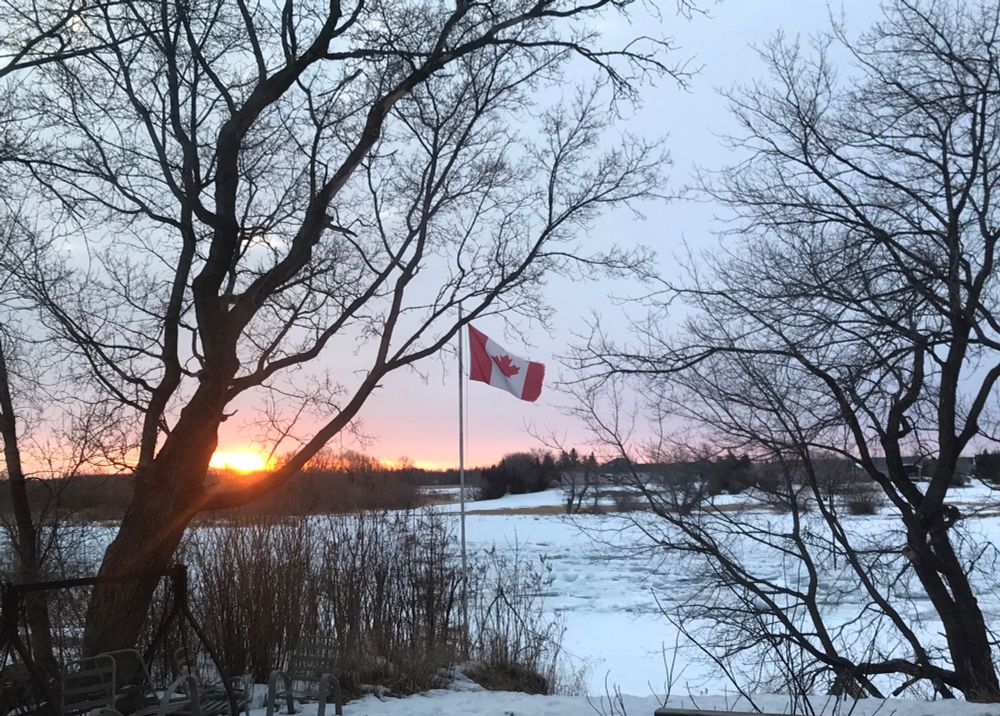 Sun rising over a winter river with a Canadian flag waving in the foreground and trees on either side 