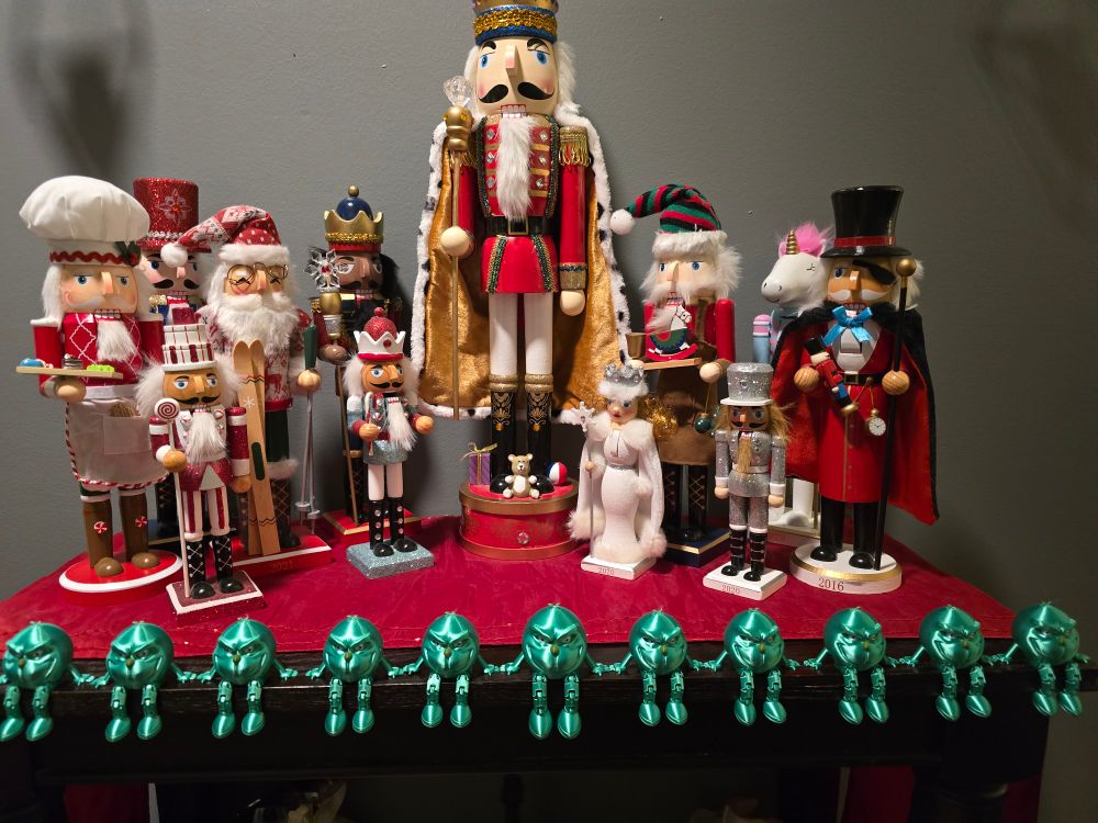 A table with red tablecloth adorned with 12 nut crackers of various shapes and sizes. Twelve 3d printed Grinch figurines are seated before them with the legs dangling over the table.